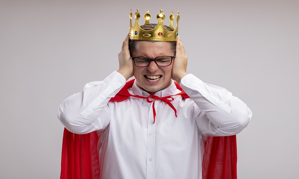super hero businessman in red cape and glasses wearing crown covering eras with hands with annoyed expression standing over white background